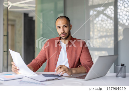 A focused businessman reviews paperwork at his desk, a laptop and calculator beside him, inside a bright office space. A focused businessman reviews paperwork at his desk, a laptop and calculator beside him, inside a bright office space. 127904929