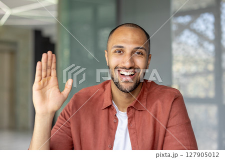 A happy man waves at the camera, smiling broadly and showing off his open palm. He's wearing a red-orange shirt. A happy man waves at the camera, smiling broadly and showing off his open palm. He's wearing a red-orange shirt. 127905012