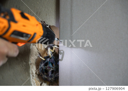 Close-up of a builder using a drill to install drywall during a home renovation, demonstrating the process of attaching building materials. Attaching a sheet of drywall to a wall using screws. Close-up of a builder using a drill to install drywall during a home renovation, demonstrating the process of attaching building materials. Attaching a sheet of drywall to a wall using screws. 127906904