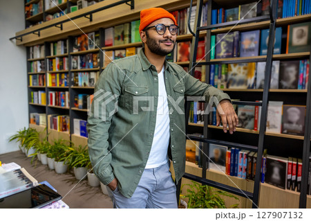 Bearded young man in an orange hat standing near the book shelf Bearded young man in an orange hat standing near the book shelf 127907132