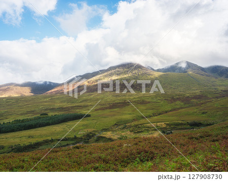 Green hills and mountains rise in the distance, partly covered by low-hanging clouds on Carrauntoohil walkway. Sunlight shines on the slopes in County Kerry, Ireland. 127908730