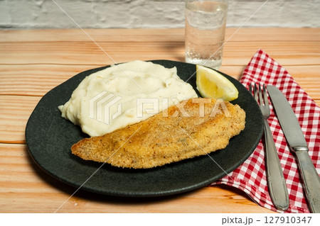 Breaded fish fillet with mashed potatoes and lemon slice on black plate 127910347