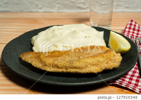 Breaded milanesa with mashed potatoes, lemon and glass of water on wooden table 127910348