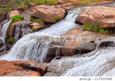 Tranquil waterfall flows over stones surrounded by lush greenery under bright sunlight. 127910486