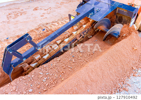 Heavy trencher equipment grinds through soil while preparing trench for underground utilities in an active construction zone 127910492