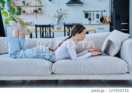 Portrait of young woman lying on sofa typing on laptop, in home interior 127910718