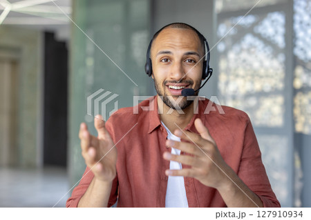 Webcam view, support worker with headset looking into camera, smiling, talking and consulting clients remotely. Man at workplace inside office Webcam view, support worker with headset looking into camera, smiling, talking and consulting clients remotely. Man at workplace inside office 127910934