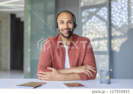 A smiling customer service representative wearing a headset in a modern office setting. He sits at a desk. A smiling customer service representative wearing a headset in a modern office setting. He sits at a desk. 127910935