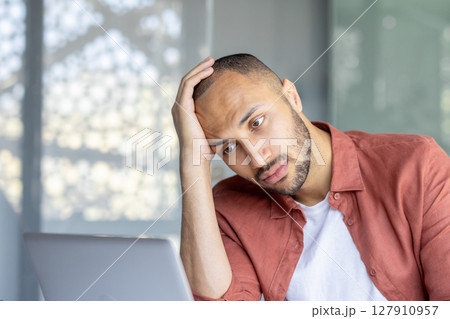 A tired man leans his head on his hand while looking at a laptop, possibly feeling stressed or overworked A tired man leans his head on his hand while looking at a laptop, possibly feeling stressed or overworked 127910957