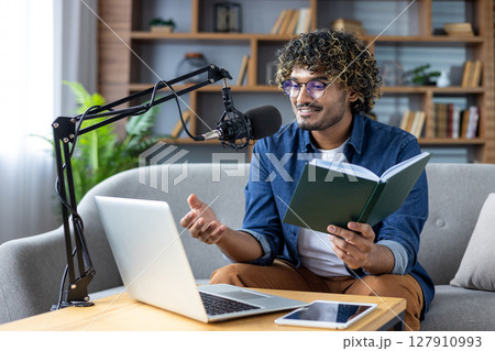 A man with glasses and curly hair records a podcast from home, holding a book and speaking into a microphone. 127910993
