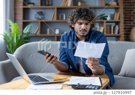 A concerned man is reviewing bills while using a laptop and a smartphone at home. Financial stress is evident in his expression. A concerned man is reviewing bills while using a laptop and a smartphone at home. Financial stress is evident in his expression. 127910997