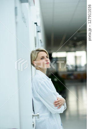 Tired female doctor leaning against wall in hospital hallway. 127911036