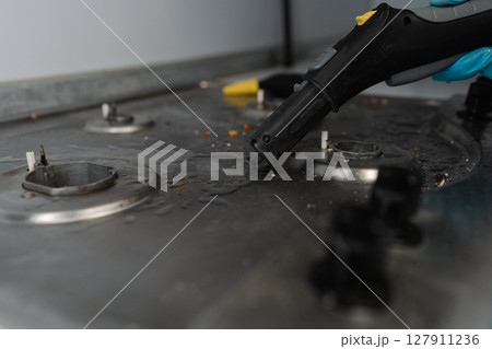 Cleaning professional wearing blue gloves using steam cleaner to remove burned-on food and grease from dirty gas stove, ensuring hygiene and cleanliness in kitchen setting, close-up. 127911236