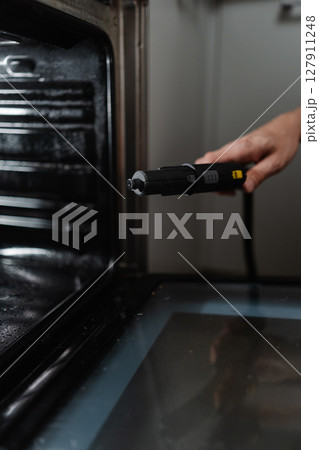 Close-up vertical shot of man sanitizing gas oven using steam cleaner, working to eliminate grime and maintain sparkling kitchen cleanliness. Concept of hygiene and cleanliness in domestic environment 127911248