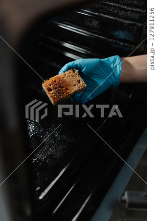 Vertical cropped shot of male hand in blue rubber glove holding dirty sponge, scrubbing soiled gas oven covered in grease stains and splashes, tackling mess in kitchen environment, close-up. Vertical cropped shot of male hand in blue rubber glove holding dirty sponge, scrubbing soiled gas oven covered in grease stains and splashes, tackling mess in kitchen environment, close-up. 127911256