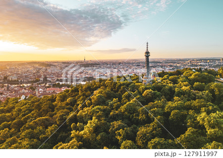 Petrin Lookout Tower rises majestically over Prague at sunrise, surrounded by lush greenery. The cityscape glows warmly as the first light of day illuminates the historic skyline. 127911997