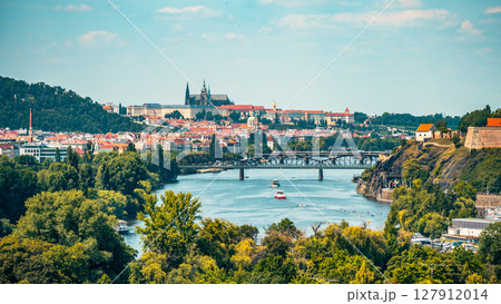 This panoramic view showcases the beauty of Prague, featuring the iconic Prague Castle and the Vltava River. Lush greenery and boats add to the charm of this historical cityscape. 127912014