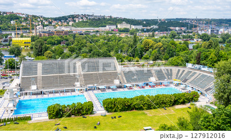 Visitors enjoy a sunny summer day at Podoli Swimming Pool in Prague. The outdoor pool area is bustling with people, surrounded by green trees and scenic views of the city. 127912016