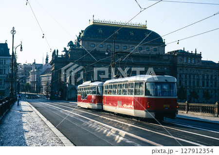Two red trams travel across a bridge near the National Theatre in Prague on a sunny morning, showcasing the vibrant public transport system in this historic city. 127912354