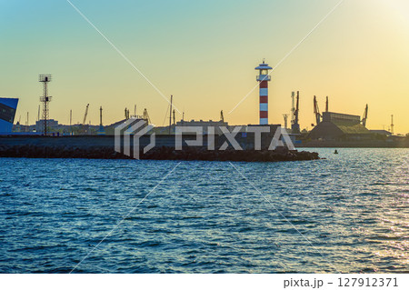 Industrial port with lighthouse at sunset. Lighthouse stands at edge of industrial port with cranes, golden sunset sky. Concept of lighthouse, urban harbor, contrast between nature and infrastructure Industrial port with lighthouse at sunset. Lighthouse stands at edge of industrial port with cranes, golden sunset sky. Concept of lighthouse, urban harbor, contrast between nature and infrastructure 127912371