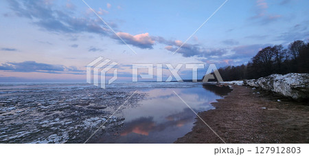 Spring landscape. Panoramic view of beautiful dawn on bay. Cumulus clouds over water in bright light. Ice, snow and rocks on coastline. Rising rays of sun are reflected in sea. High quality photo 127912803