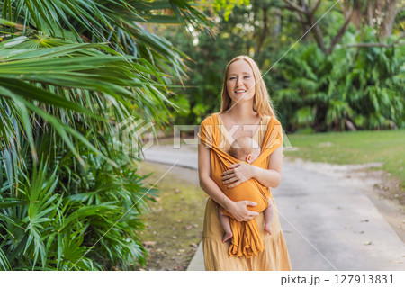 Mother holding her little baby in a yellow sling in the park. Warm and loving family moment. Babywearing, parenting, and mother-child bonding concept 127913831