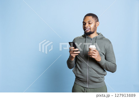 Confident relaxed person texting his friends and serving fresh cup of coffee, feeling happy and smiling at his smartphone. African american lively man messaging his pals, drink caffeine. Confident relaxed person texting his friends and serving fresh cup of coffee, feeling happy and smiling at his smartphone. African american lively man messaging his pals, drink caffeine. 127913889