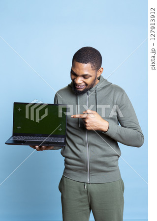 Confident happy person pointing at green screen mockup in studio, using personal laptop for an advertisement on camera. African american young guy gesturing against blue background. Confident happy person pointing at green screen mockup in studio, using personal laptop for an advertisement on camera. African american young guy gesturing against blue background. 127913912