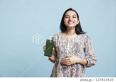 Indian woman showing isolated copy space display in front of the camera, creating an advertisement for a commercial against blue background. Young joyful adult holding device in studio. 127913919