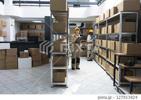Woman employee conducting manual labor surrounded by packages and crates, staff managing shipping, delivery and inventory control tasks. Warehouse interior supply and demand operations. 127913924