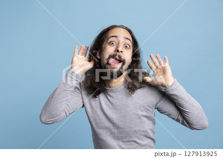 Playful young man with a long hair and beard making a goofy face in the studio. Carefree guy radiates humor and joy, capturing a lighthearted moment full of laughter and casual personality. Playful young man with a long hair and beard making a goofy face in the studio. Carefree guy radiates humor and joy, capturing a lighthearted moment full of laughter and casual personality. 127913925