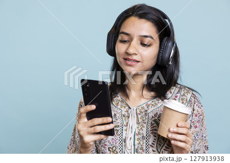 Pleased excited woman playing music on phone and drinking coffee, having fun dancing on her favorite songs against blue background. Delighted indian girl wearing headphones and doing funky moves. Pleased excited woman playing music on phone and drinking coffee, having fun dancing on her favorite songs against blue background. Delighted indian girl wearing headphones and doing funky moves. 127913938