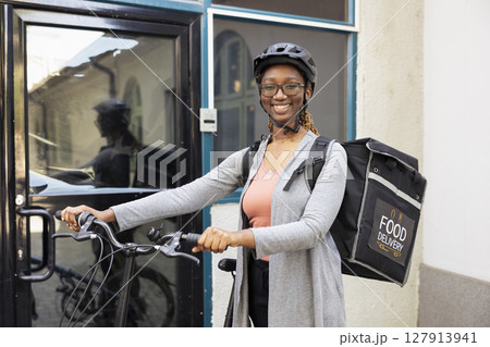 Portrait of smiling black woman working as a takeaway courier on the bike, riding with a helmet and carrying her backpack. Takeout employee delivering fast food or other meal orders. Portrait of smiling black woman working as a takeaway courier on the bike, riding with a helmet and carrying her backpack. Takeout employee delivering fast food or other meal orders. 127913941