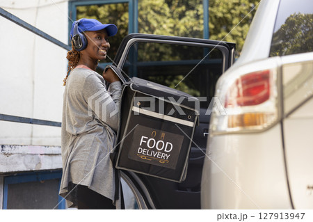 African american woman delivering takeout with the car using a backpack, arriving at the entrance of a modern building. Preparing to ring the door bell to bring the food order to client. 127913947
