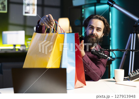 Media star holding colorful shopping bags in hand, showing fans purchases he recently got, filming himself in apartment. Content creator in studio presenting audience new acquisitions 127913948
