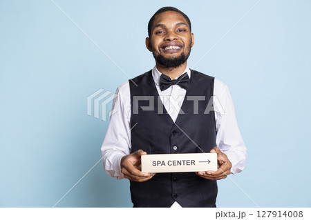 Hotel porter in suit and tie helping with directions to the spa center using a pointer, holding the wall sign to guide guests to resort amenities. Providing great concierge services. 127914008