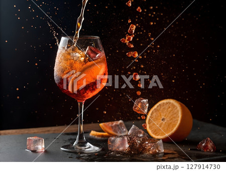 Aperol Spritz being poured into a wine glass with splashes and motion freeze, floating ice cubes and orange slice 127914730