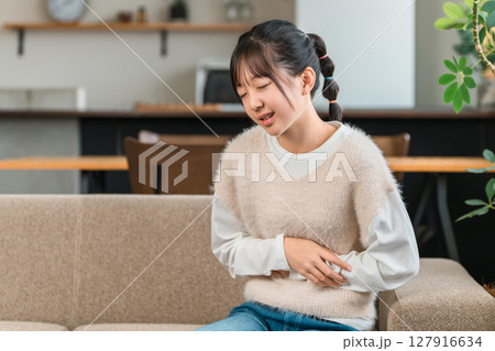 自宅のソファに座り腹痛でお腹を抑える小学生,中学生の女の子 127916634