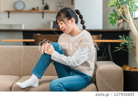 自宅のソファに座り膝痛で膝を抑える小学生,中学生の女の子 127916885