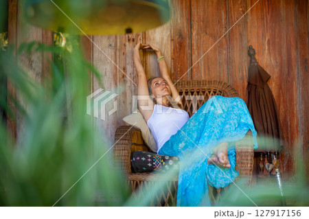 Young woman sit on open-air veranda of wooden bungalow with tropical garden view. Girl relaxing in luxury villa on family summer vacation. Countryside lifestyle. 127917156
