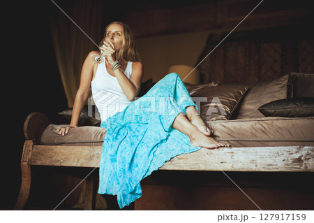Young woman sit on sofa inside luxury wooden villa. Girl relaxing on family summer vacation. Tropical lifestyle. 127917159