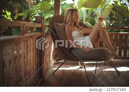 Young woman sit on open-air veranda of wooden bungalow with tropical garden view. Girl relaxing in luxury villa on family summer vacation. Tropical lifestyle. Young woman sit on open-air veranda of wooden bungalow with tropical garden view. Girl relaxing in luxury villa on family summer vacation. Tropical lifestyle. 127917160