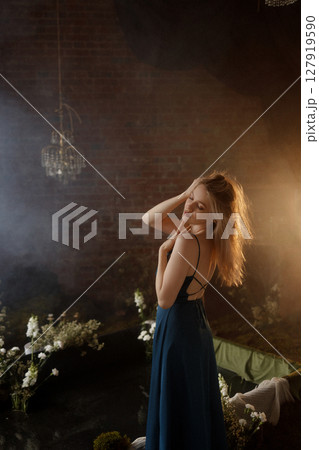 Woman in blue dress posing under vintage chandelier in dark room with brick wall and flowers 127919590