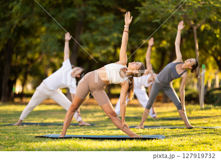 Yoga lesson visitors with coach perform Trikonasana in park 127919732
