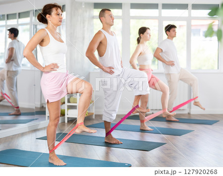 Young men and women practicing hip workout with band standing on mat 127920268