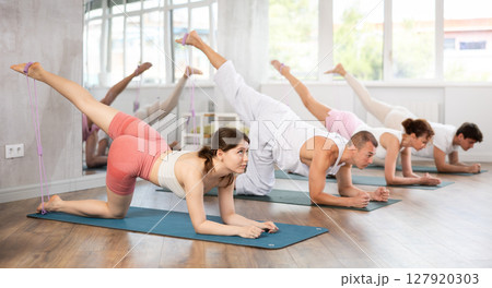 Happy girl on hands and knees doing stretch exercises for legs with resistance band on mats with group of sporty people in gym Happy girl on hands and knees doing stretch exercises for legs with resistance band on mats with group of sporty people in gym 127920303