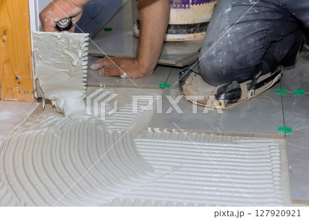 Worker is spreading tile adhesive on floor in home renovation project, preparing for tile installation. 127920921