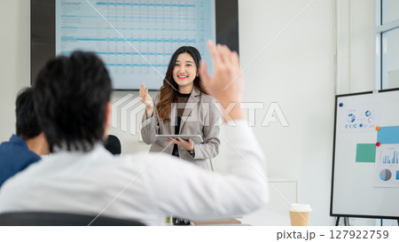 Businesswoman or female office worker is looking at male coworker who raised a hand to ask question. 127922759