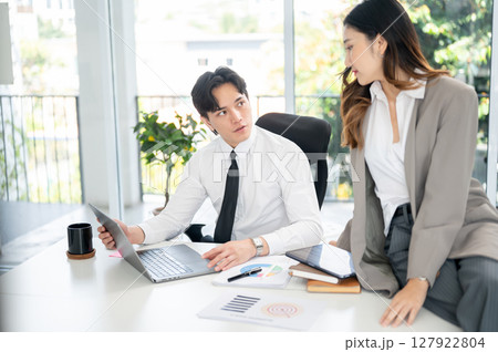 Asian male office worker is asking or talking with female coworker about the data on laptop screen. 127922804