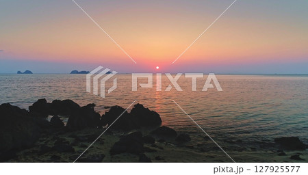 Sun setting over calm sea creating colorful sky reflecting on water surface, with dark rocks and beach in the foreground, Koh Samui, Thailand 127925577